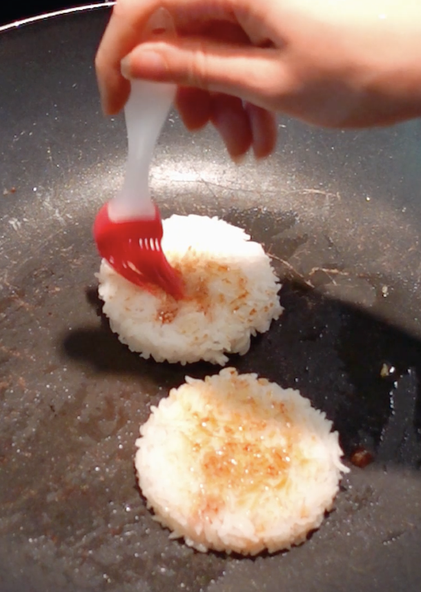 Hand brushing soy sauce on rice patties in a pan for a crispy rice burger recipe